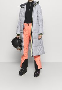 Personne portant un long manteau d'hiver gris, un pantalon de ski corail avec des patchs noirs, des bottes de ski noires, tenant un casque noir.