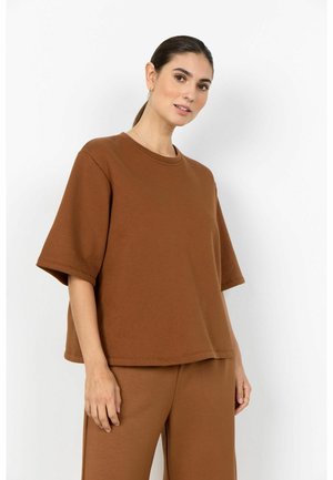 Kvinde med mørkt hår i en lav hestehale iført en løs brun kortærmet top og matchende brune bukser, stående mod en hvid baggrund.