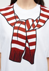 Λευκό μπλουζάκι με γραφικό μοτίβο κόκκινης και λευκής ριγωτής κασκόλ δεμένης γύρω από την περιοχή του λαιμού, φορεμένο από ένα άτομο με σκούρα μαλλιά.