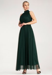 Długa zielona sukienka z unoszącym się materiałem, z wysokim kołnierzem i marszczonym biustem, posiada przezroczysty overlay i rozkloszowany krój.