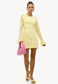 Vestido mini amarillo de encaje con mangas largas de campana, combinado con un bolso rosa y unos tacones amarillos de punta abierta. Patrón intrincado con bordes en forma de concha.