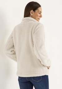 Cremfarbene Fleecejacke mit hohem Kragen, langen Ärmeln und seitlichen Taschen. Weicher Stoff mit leicht übergroßem Schnitt.