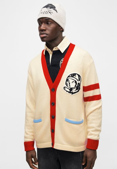 Κρεμ ζακέτα με κόκκινες λεπτομέρειες, μπλε τσέπες και μαύρο σχέδιο αστροναύτη. Φορεμένη πάνω από μαύρο πουκάμισο με γιακά, με μπεζ σκούφο.