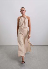 Top halter beige con escote recogido, combinado con una falda larga y fluida que presenta un detalle de lazo. Usado con sandalias marrones de tiras.