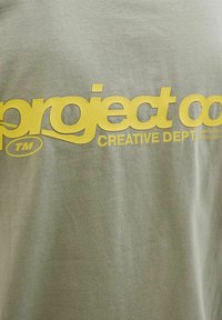 Lyse olivengrøn bomulds t-shirt med iøjnefaldende gul grafisk tekst og logo-design, med en glat tekstur og afslappet pasform.