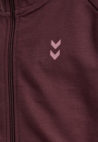 Burgundy zip-jacka i mjukt tyg; har en rosa triangulär logotyp och synliga sömmar längs dragkedjan och nederkanten.