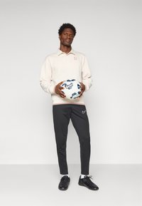 Mand, der står med en hvid og blå Nike fodbold, iført en cremfarvet sweatshirt, sorte Nike bukser, hvide sokker og sorte sko.