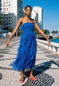 Femme aux cheveux tressés, vêtue d'une robe bustier en tulle bleu et de tongs roses, debout sur une promenade en bord de mer à motifs sous un ciel clair.