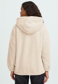 Beige fliiskangaga kapuutsiga pusa, millel on pehme, tekstuuriga pind, suur kapuuts ja lõdva lõikega, koos kogutud varrukate ja sirge äärisega.