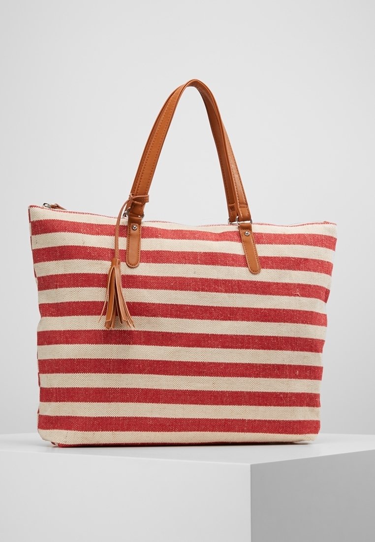 Sac cabas rayé rouge et beige avec anses en cuir fauve et un détail pompon en cuir, posé sur un piédestal blanc.