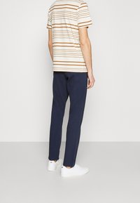 Randig t-shirt i beige med bruna ränder, i kombination med marinblå byxor. Person som bär vita sneakers, sedd bakifrån.