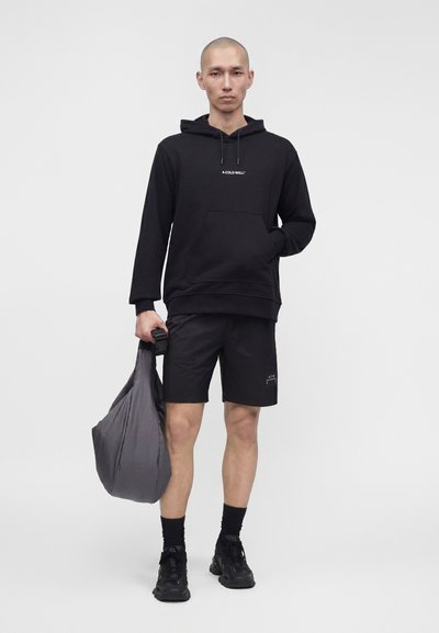 Άνδρας μοντέλο που φοράει μαύρο φούτερ και σορτς A-COLD-WALL, κρατώντας μεγάλη γκρι τσάντα, στέκεται μπροστά σε απλό ανοιχτό φόντο.