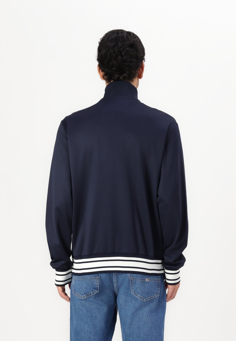 Chaqueta bomber de color navy con alto cuello, puños y dobladillo de rayas blancas, hecha de un tejido suave, mostrada desde la parte trasera en una persona que lleva vaqueros.