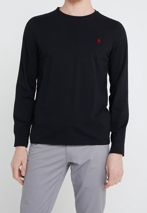 Homme portant un t-shirt noir à manches longues avec un petit logo rouge sur le côté gauche de la poitrine, associé à un pantalon gris clair.