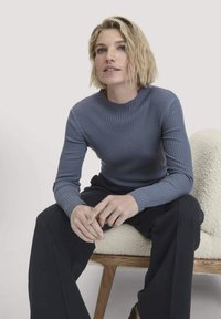 Junge Frau mit kurzem blonden Haar, die einen blauen, gerippten Langarmpullover und schwarze Hosen trägt und auf einem strukturierten weißen Stuhl sitzt.