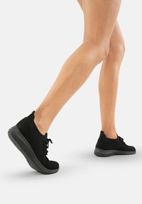 Zapatillas ligeras negras con parte superior de tejido texturizado, suela acolchada ycordones redondos. El diseño presenta un corte bajo con una suela flexible.