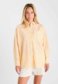 Femme portant une chemise à boutons rayée verticale jaune et blanche, avec une poche poitrine rayée, et un short blanc, se tenant devant un fond uni.