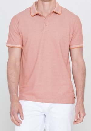 Man draagt een zalmroze poloshirt met korte mouwen, een oranje gerande kraag en witte broek, staand tegen een effen witte achtergrond.