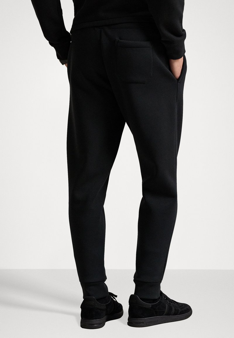 Svarte joggingbukser med en tætsiddende pasform, elastisk talje og sidelommer. Har en baglomme og ribbede ankelmanchetter. Båret med sorte sko.