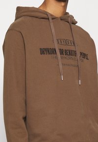 Sudadera marrón con cordón, que presenta un tejido texturizado y un logo en texto impreso en negro en la parte delantera. Corte regular y mangas largas.