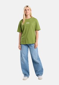 Camiseta de algodón verde con mangas cortas y un corte relajado, con un pequeño gráfico blanco en el pecho. Combinada con jeans de pierna ancha de color azul claro.