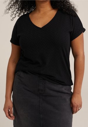 Mujer con una blusa negra texturizada de manga corta y cuello en V, metida dentro de una falda vaquera oscura de talle alto con costuras frontales.