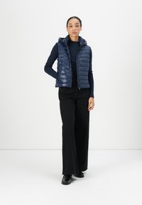 Navy puffer vest with a hood, quilted design, zip closure, worn over a fitted long-sleeve top, paired with black wide-leg pants and shoes.