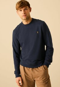 Navy blauwe trui gemaakt van zacht materiaal met lange mouwen, ronde hals en een klein gouden logo op de borst, gecombineerd met beige broek.