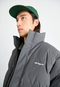 Chaqueta acolchada gris con un alto cuello, que presenta un logo de Carhartt. Lleva una camisa verde debajo y se complementa con una gorra de béisbol verde.