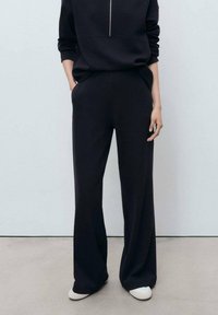 Personne portant un pantalon large noir, un haut à manches longues noir avec fermeture éclair, les mains dans les poches et des chaussures blanches se tenant sur un sol gris.