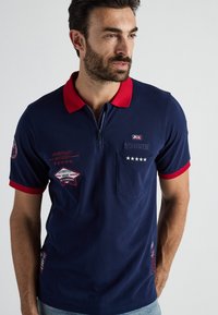 Granatowa koszulka polo z czerwonym kołnierzykiem, zamkiem błyskawicznym i krótkimi rękawami. Posiada haftowane detale, w tym kieszonkę i akcenty logo.