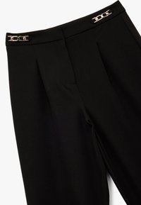Pantalones negros a medida con pliegues, bolsillos laterales y acentos en tono dorado en la cinturilla. Textura suave, acabado de tela lisa.