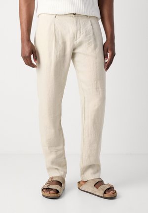 Man die beige linnen broek en beige suède sandalen draagt, staand tegen een witte achtergrond.