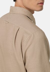 Beige, textuurshirt met een klassieke kraag. Heeft subtiele horizontale strepen en een enkele knoop op de achterkant van de kraag. Getailleerde pasvorm.