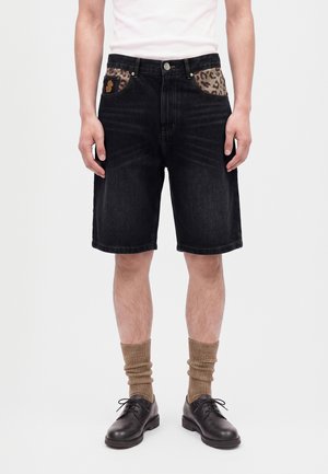 Homme portant un short en jean noir avec une doublure de poche imprimée léopard, des chaussettes marron, des chaussures habillées noires et une chemise blanche, debout devant un fond uni.