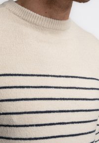 Light beige sweater with a ribbed crew neckline, featuring navy horizontal stripes and a soft, textured fabric. Close-up view.
