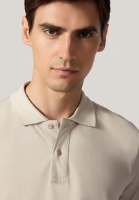Beżowa koszulka polo z teksturowanym materiałem, z dwoma guzikami przy kołnierzy i klasycznym kołnierzykiem. Zbliżenie na obszar klatki piersiowej.