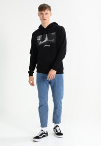 Black hooded sweatshirt featuring a graphic of hands in prayer and text "pray," paired with light blue jeans and black sneakers.