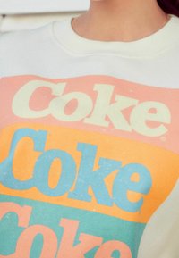 White cotton sweat shirt featuring retro multi-colored "Coke" text in pink, orange, and blue. The design includes a slightly faded texture.