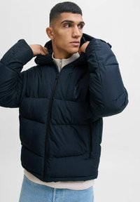 Chaqueta puffa azul marino con cierre frontal, capucha y puños elásticos. Presenta un diseño acolchado y dos bolsillos laterales con cremalleras.