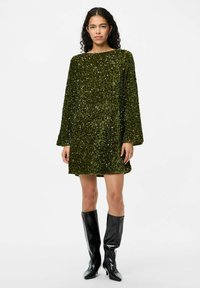 Zaļš spīdīgs mini kleita ar garām piedurknēm, brīvu piegriezumu un laivu kakla izgriezumu, apvienota ar spīdīgām melnām augstpapēžu zābakām līdz ceļgaliem.