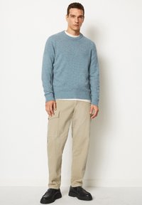 Hellblauer Strickpullover mit Rundhalsausschnitt, kombiniert mit beigen Cargo-Hosen mit Taschen. Schwarze Schnürschuhe runden das Outfit ab.