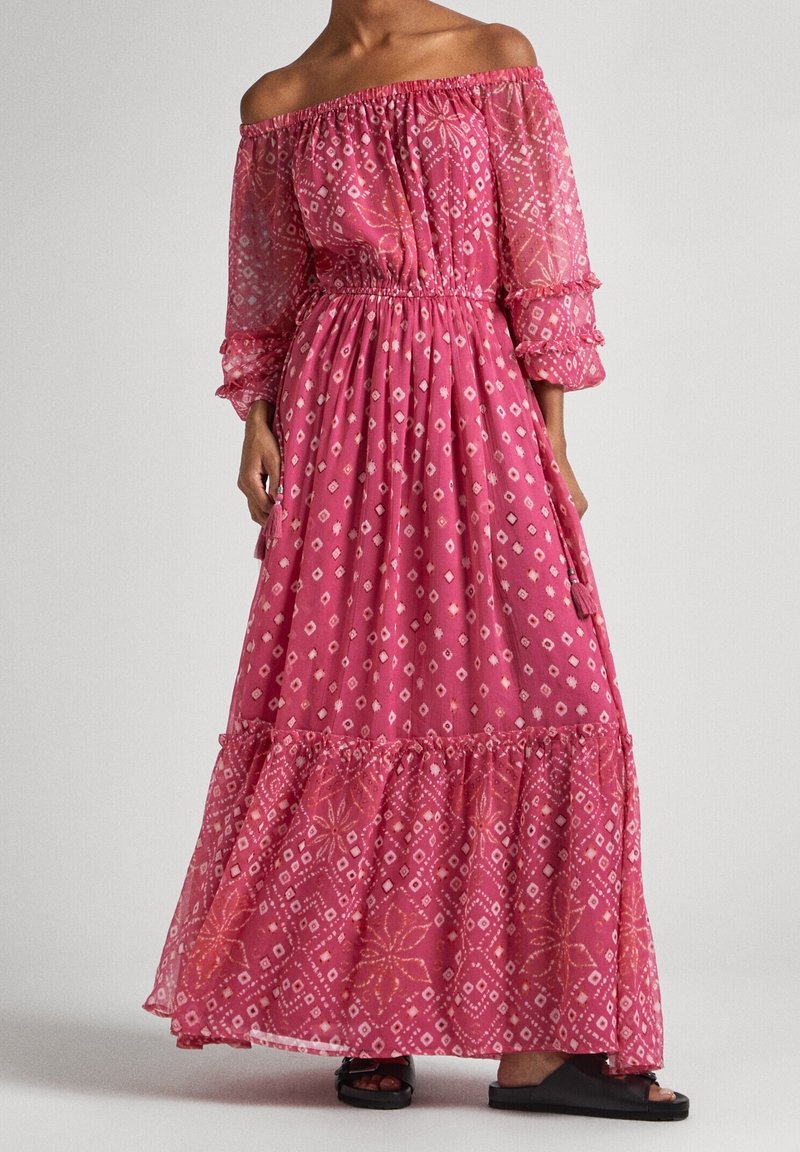 Femme portant une longue robe rose à épaules dénudées avec des motifs géométriques blancs et un ourlet à volants, associée à des sandales noires à bouts ouverts.