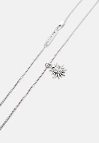 Collier en chaîne argentée avec un pendentif en forme de soleil présentant des rayons texturés et un fermoir mousqueton avec une petite étiquette rectangulaire.