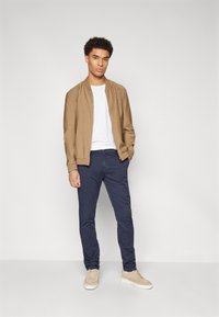 Giacca bomber marrone sopra una t-shirt bianca, abbinata a pantaloni chino blu navy e sneakers beige. Design semplice, vestibilità comoda e stile informale.