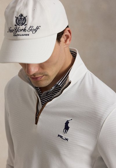 Άνδρας που φοράει λευκό καπέλο New York Golf Ralph Lauren και λευκή μπλούζα με φερμουάρ, με γιακά με ναυτικό ρίγες και λογότυπο παίκτη πόλο.