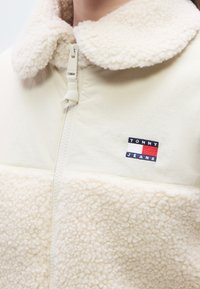 Μπεζ ζακέτα φλις με γιακά, μπροστινό φερμουάρ μέχρι τη μέση και κεντημένο λογότυπο στο στήθος. Μαλα κή υφή και ζεστό σχέδιο.