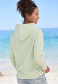 Felpa verde chiaro con una texture morbida, maniche lunghe e cappuccio con cordini. Abbinata a jeans azzurri chiari con un dettaglio del logo.