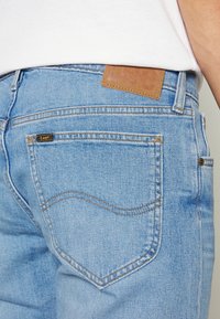 Světle modré džíny z denimu s klasickým střihem, vybavené zadními kapsami, hnědým koženým štítkem v pase a viditelným oranžovým prošíváním.