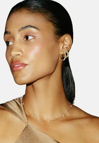 Gouden creolen met heldere accenten, delicate gouden ketting met kleine kralen, en een gladde satijnen bruine top. Close-up van een profiel.